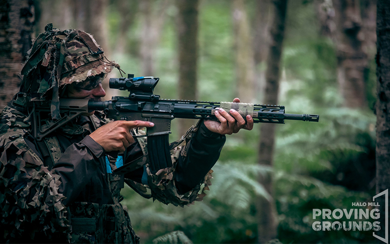 An Airsofter aiming down an optic in the woods.